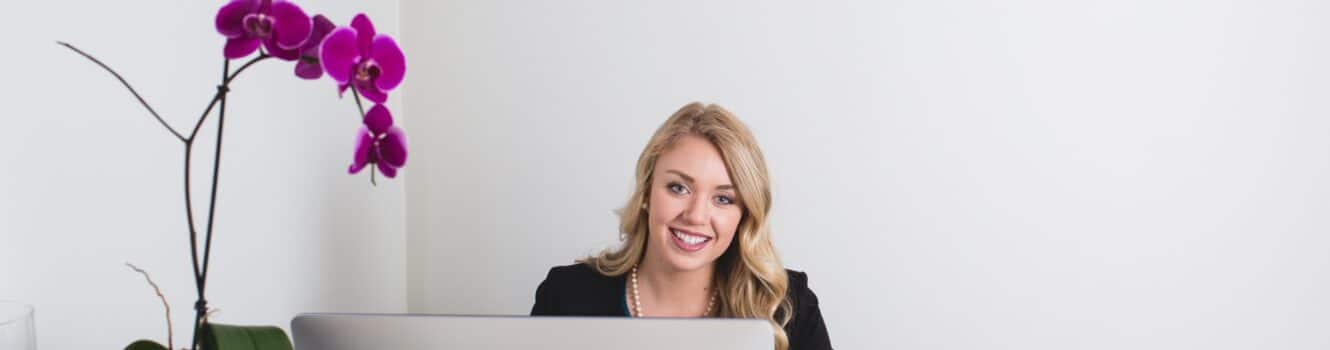 Woman smiling sitting behind a desk with an orchid on it.